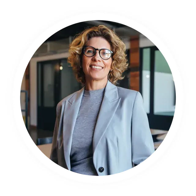 A smiling woman with curly hair and glasses, wearing a light grey blazer in an office